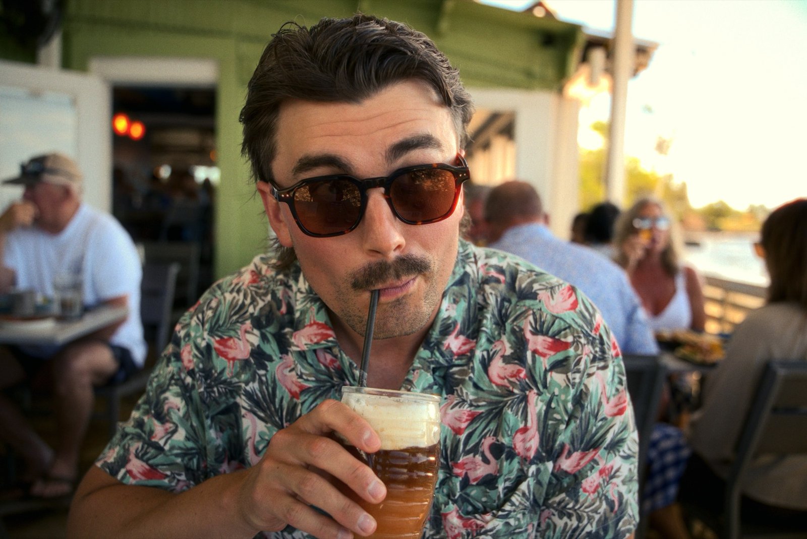 Eric Hunsicker in a flamingo shirt, sunglasses on, sipping a cold brew at a waterfront patio